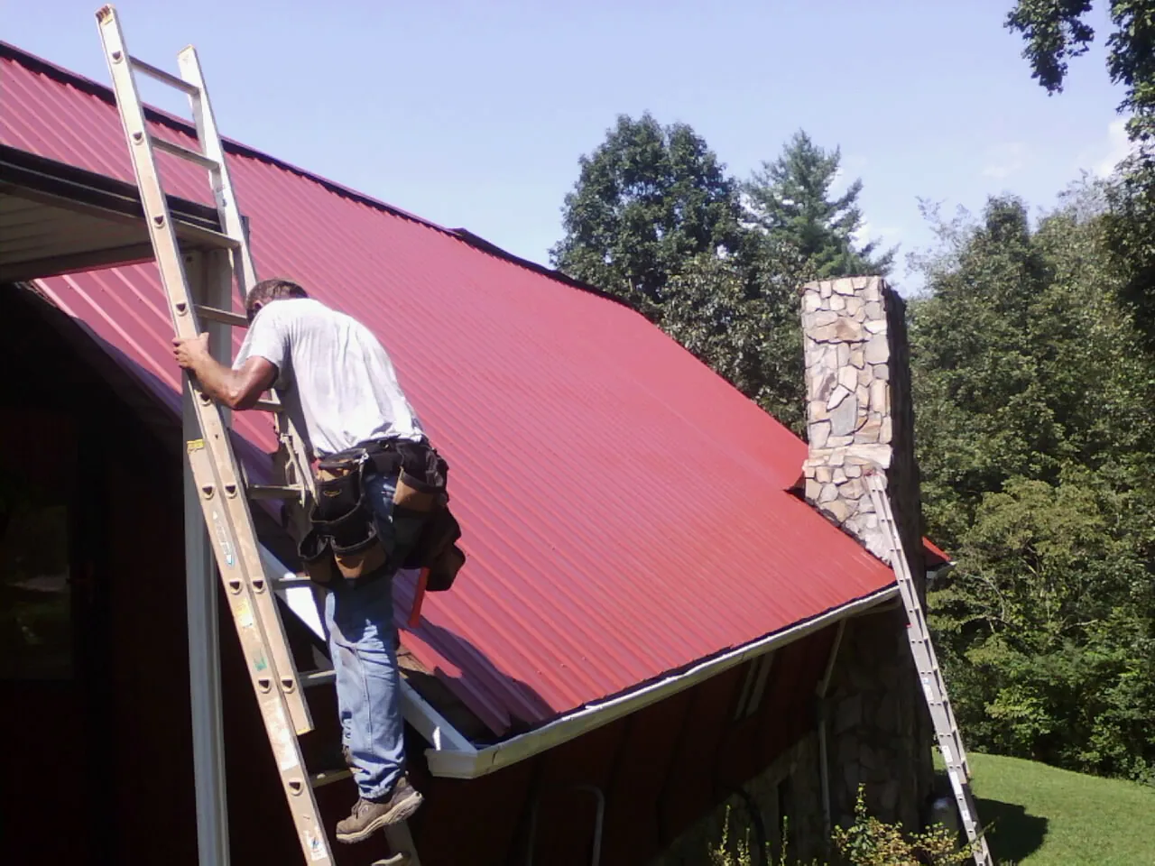 Roofer on a ladder beside a red metal roof and stone chimney — Soffit & Fascia Repair by Roofing Brothers in Maple Park